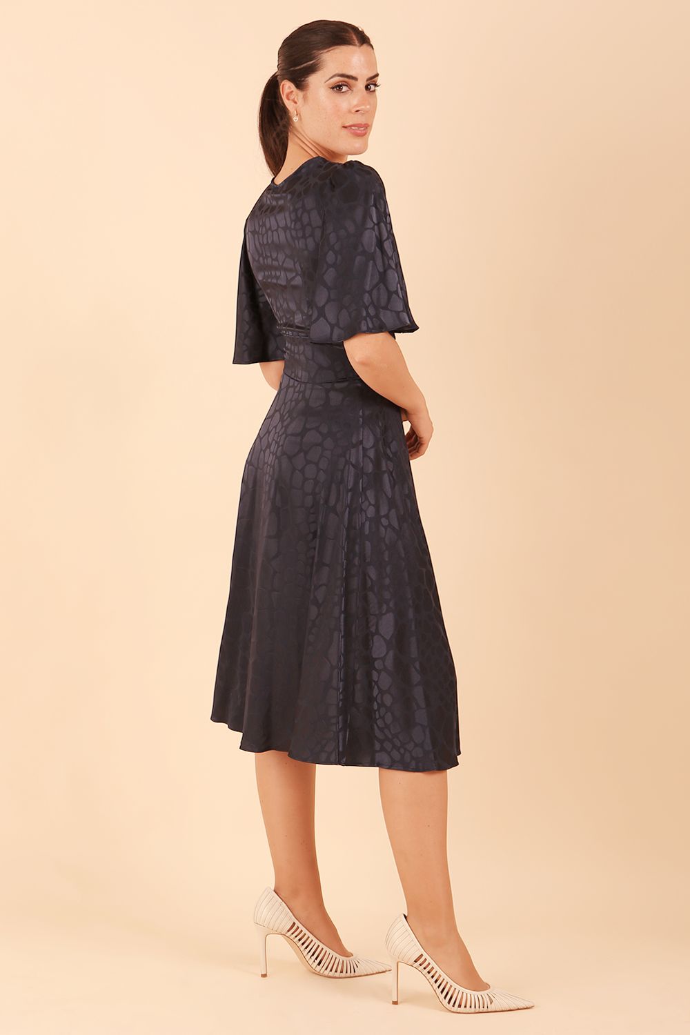 brunette model wearing diva catwalk cambrian satin swing dress with bell short sleeve wide asymmetric waistband and pleating under bust and low v-neckline in navy back