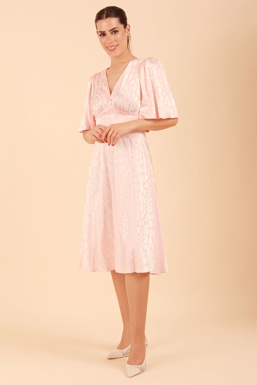 brunette model wearing diva catwalk cambrian satin swing dress with bell short sleeve wide asymmetric waistband and pleating under bust and low v-neckline in pink front