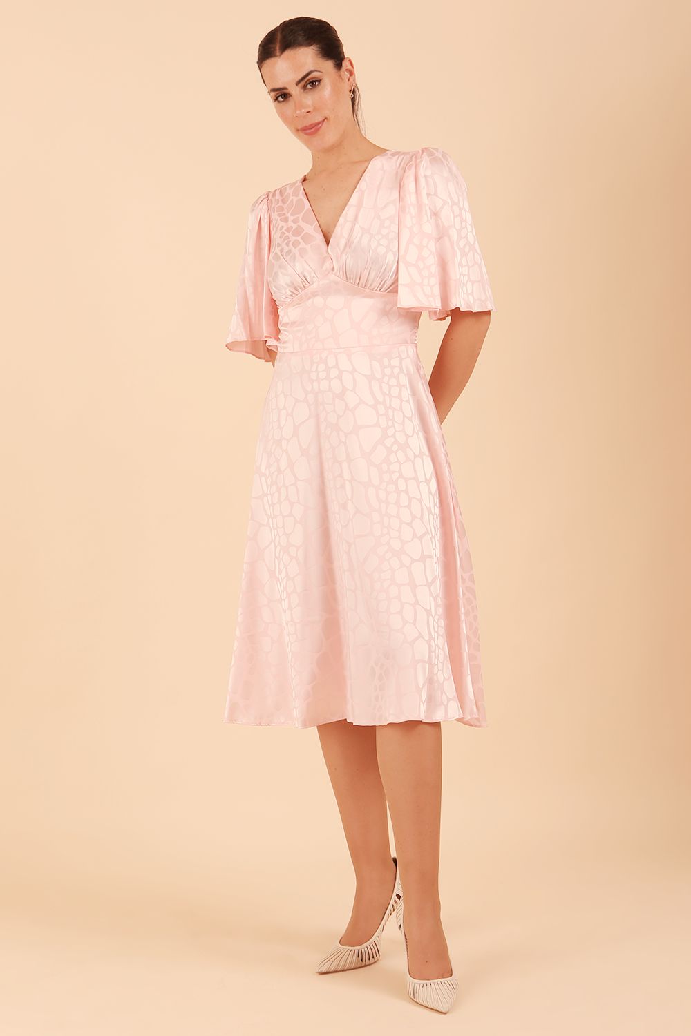 brunette model wearing diva catwalk cambrian satin swing dress with bell short sleeve wide asymmetric waistband and pleating under bust and low v-neckline in pink front